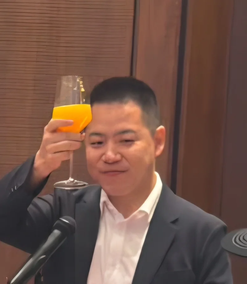 A man in a dark suit and white shirt, identified as Gao Jin, is seen raising a glass filled with a yellow-orange beverage in a toast. He is standing behind a microphone in a room with wood-paneled walls, suggesting a formal business or diplomatic event.