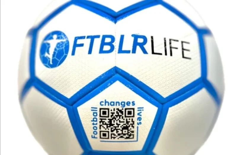 A close-up photograph of a white and blue football featuring the FTBLRLIFE logo and a circular icon of a soccer player. Below the main branding, a QR code is visible surrounded by the slogan 'Football changes lives'.