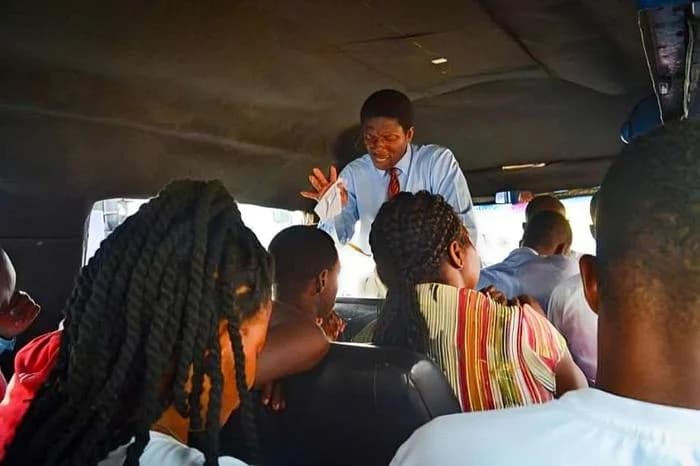 A man dressed in a light blue shirt and red tie stands at the front of a crowded commercial transport vehicle, gesturing while apparently preaching to passengers. The interior view from the back shows several passengers seated and facing him, capturing a common religious practice in Ghanaian public transport.