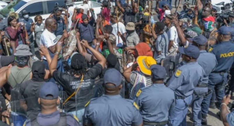 A group of protesters, some carrying traditional shields and sticks, confront a line of South African police officers in blue uniforms. The scene depicts a tense standoff during a public demonstration, with civilians raising their hands and several people documenting the event on mobile devices.