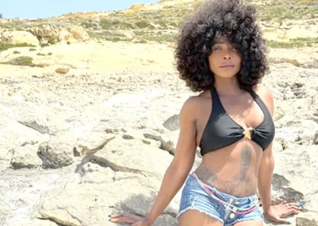 Ghanaian-British singer Stephanie Benson is pictured posing on a rocky coastline. She is wearing a black bikini top, denim shorts, and waist beads, with her signature voluminous curly hair framing her face.