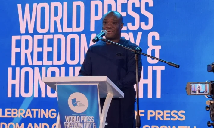 Felix Kwakye Ofosu, a prominent Ghanaian politician, is captured speaking at a podium during a World Press Freedom Day and Honours Night event. He is wearing a dark blue traditional outfit and stands before a large blue backdrop with white lettering emphasizing media freedom.