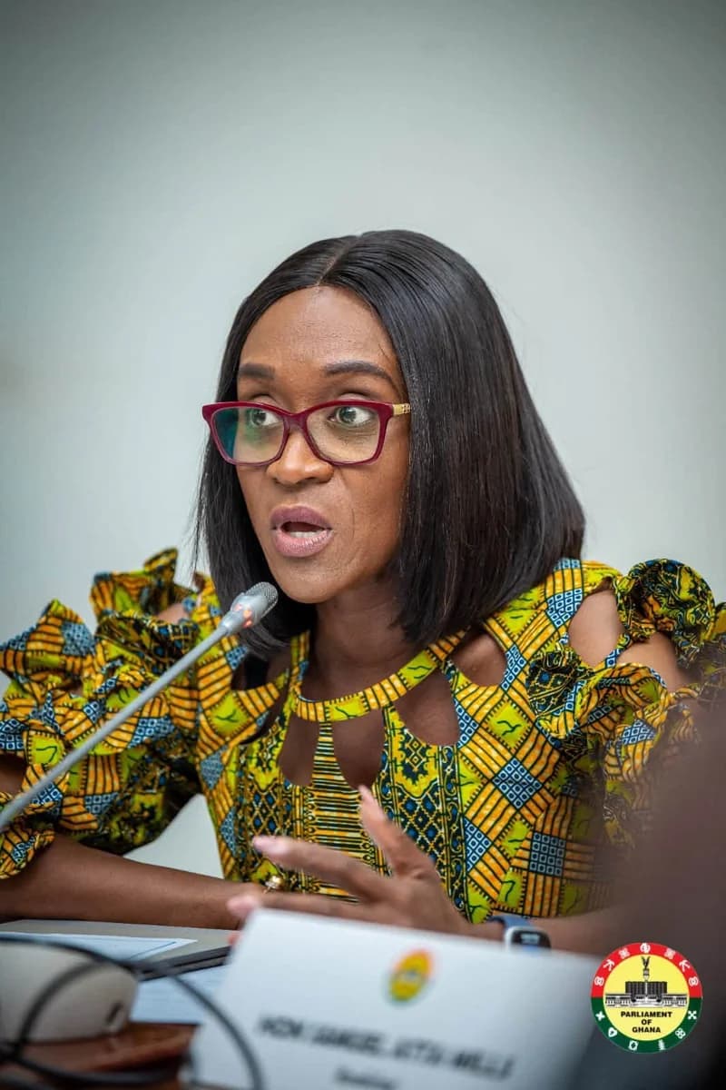 A woman wearing red-framed glasses and a vibrant yellow and green patterned dress is speaking into a microphone during an official proceeding. A nameplate for Hon. Samuel Atta-Mills and the official Parliament of Ghana logo are visible in the foreground, suggesting a legislative setting.