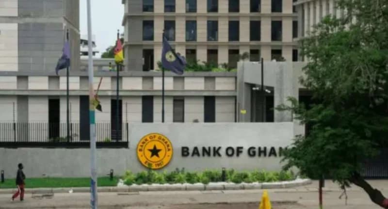 An exterior view of the Bank of Ghana building in Accra, featuring the central bank's logo and name prominently on a perimeter wall. Flags are visible flying in front of the modern multi-story office complex, while a lone pedestrian walks past the entrance.