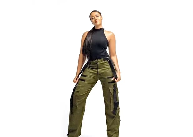 A professional studio photograph of Nigerian actress Damilola Adegbite posing against a plain white background. She is wearing a black sleeveless turtleneck top paired with olive green cargo pants that feature distinctive black strap detailing.
