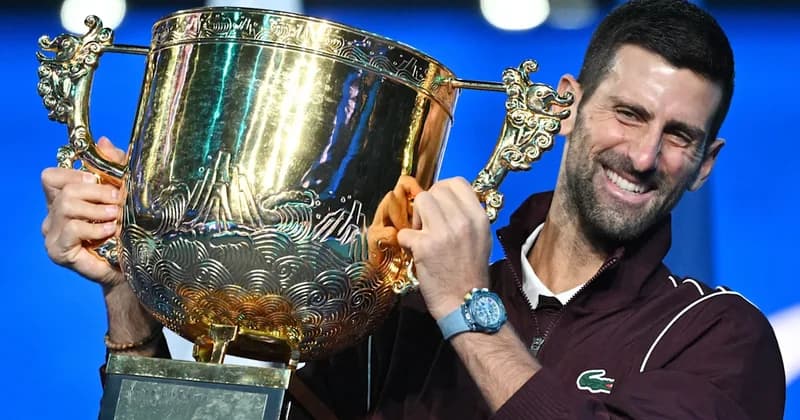 Tennis star Novak Djokovic is pictured smiling broadly while holding a large, ornate gold trophy following a major tournament victory. He is wearing a maroon Lacoste-branded jacket and a light blue wristwatch against a vibrant blue background.