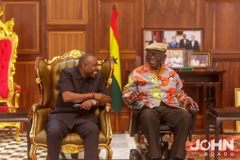 Former Ghana President John Kufuor and former NPP General Secretary John Boadu are captured in a shared moment during a courtesy visit. They are seated in an elegantly wood-paneled room featuring the Ghana flag and several framed historical photographs in the background.