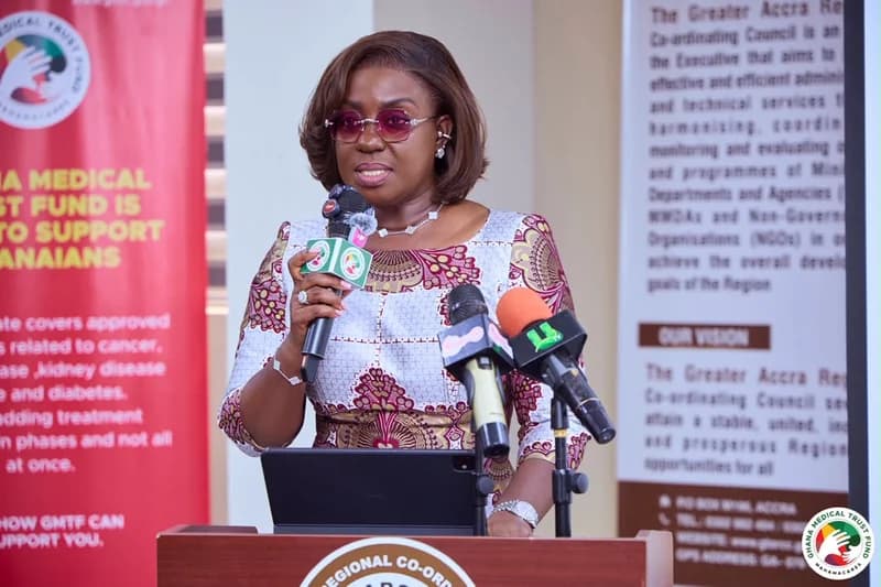 A woman in a vibrant patterned dress and tinted glasses is captured delivering a speech at a podium. She is surrounded by microphones and flanked by banners for the Ghana Medical Trust Fund and the Greater Accra Regional Co-ordinating Council.