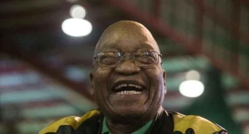 A close-up portrait of former South African President Jacob Zuma laughing. He is wearing his signature spectacles and a jacket with yellow and black patterns, likely in a public or political setting with blurred indoor lighting in the background.