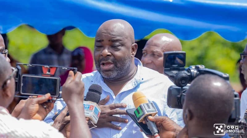 The image shows Michael Okyere Baafi, Member of Parliament for New Juaben South, addressing media personnel at an outdoor event. He is surrounded by various microphones and cameras while speaking under a blue canopy.