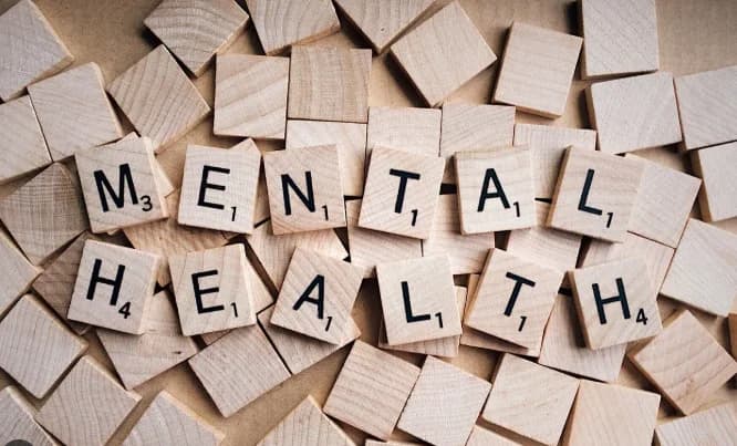 Wooden letter tiles are arranged to spell out the words 'MENTAL HEALTH' against a background of scattered blank wooden tiles. The image uses a clean, top-down perspective to highlight the central message regarding psychological well-being.