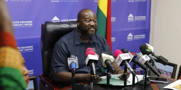 Kwabena Ampofo Appiah, Managing Director of the State Housing Company (SHC), addresses a press conference in Ghana. He is seated behind a desk crowded with microphones from various media outlets, positioned in front of an SHC-branded backdrop and the national flag.