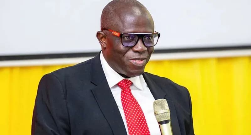 Justice Emmanuel Yonny Kulendi, a Supreme Court Justice of Ghana, is captured speaking into a microphone during an event. He is wearing a dark suit, a white shirt, and a red polka-dot tie against a backdrop of yellow drapery and a white screen.