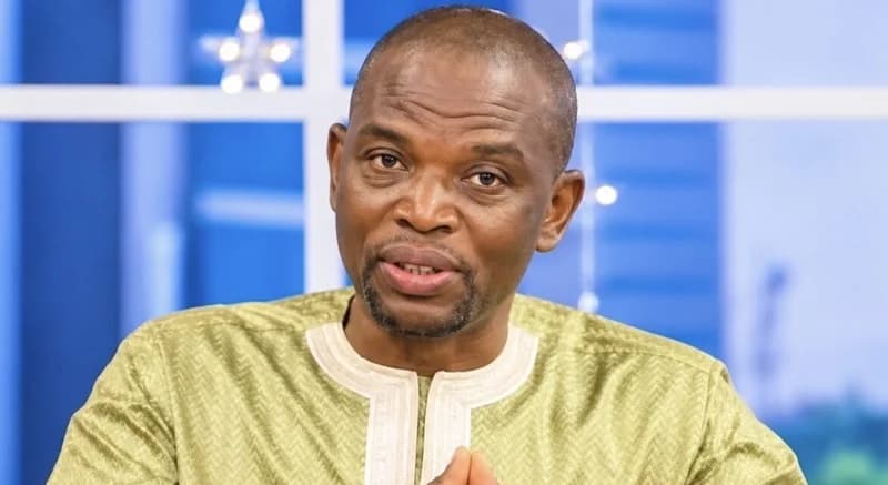 A close-up photograph of a man, identified in the context as Bataglia, wearing a light green traditional Ghanaian smock. He is looking directly at the camera with a focused expression against a blue and white studio backdrop.