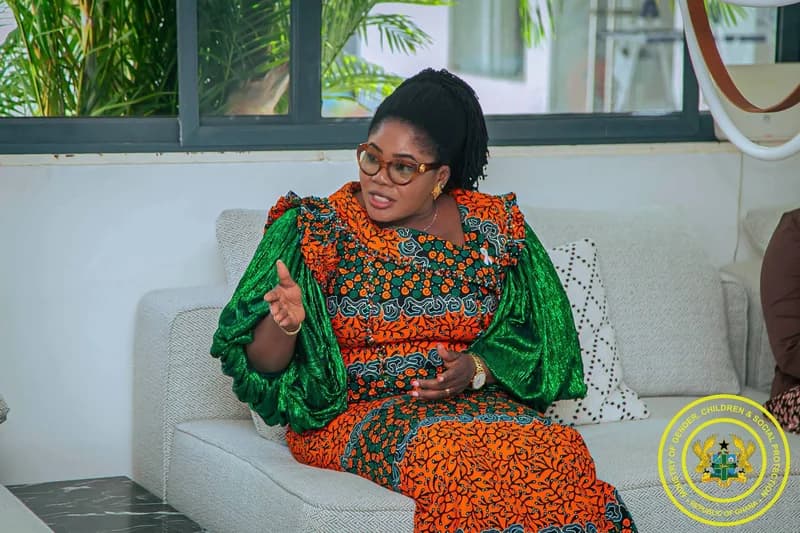 A woman, likely a government official, is captured speaking while seated on a light-grey sofa. She is wearing a vibrant orange and green African print dress with voluminous green satin sleeves and the official seal of the Ministry of Gender, Children and Social Protection is visible in the bottom right corner.