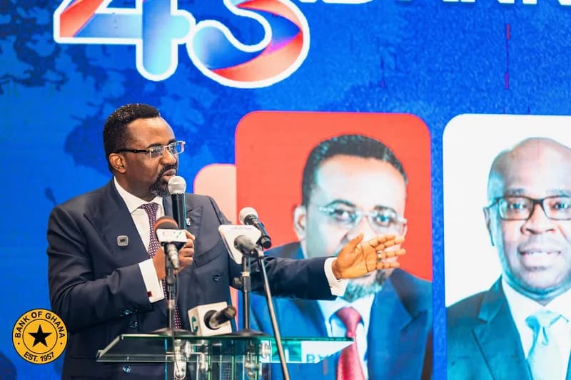 A gentleman in a dark suit and glasses is seen speaking at a podium with multiple microphones during a formal event. The background features a large blue screen with a '43rd' anniversary graphic and the Bank of Ghana logo, indicating an official financial sector ceremony or announcement.
