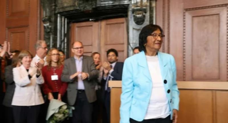 Navi Pillay, a prominent jurist and former UN High Commissioner for Human Rights, walks through a hall while a group of people applauds. She is seen wearing a light blue blazer and glasses in what appears to be a formal international setting or courtroom.