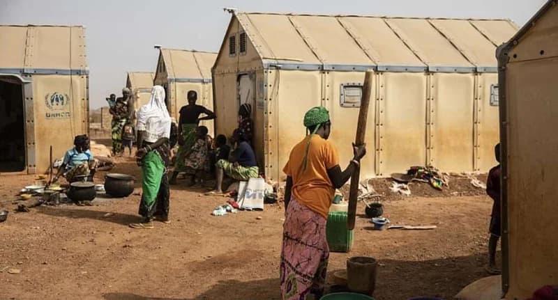 This photograph shows an internally displaced persons camp in the Sahel, with several tan-colored UNHCR shelters lined up. Women and children are engaged in daily activities such as cooking over open fires and pounding food, illustrating the humanitarian conditions in the region.