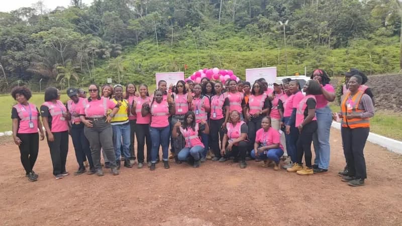 A large group of women, likely employees of Adamus Resources, are pictured outdoors wearing pink t-shirts and pink reflective safety vests for a breast cancer awareness event. They are posed in front of a hilly, forested background with pink balloons and banners visible behind the group.