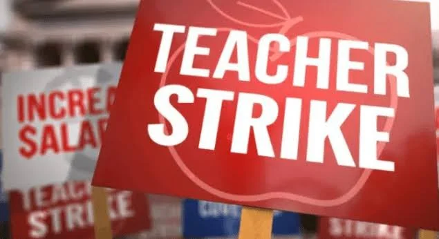 A close-up photograph of protest placards held during a labor demonstration. The main red sign features the text 'TEACHER STRIKE' in bold white capital letters over an apple graphic, while background signs mention salary increases.
