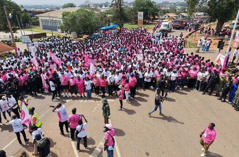 A large crowd of participants, many dressed in pink and white t-shirts, gathers for a breast cancer awareness walk in Ghana. The aerial-style shot shows a sea of people carrying banners and signs through a town street, with an ambulance and fire truck visible in the background.