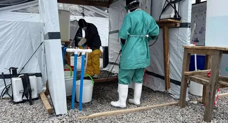 Healthcare workers in protective gear operate at a sanitation station within a temporary medical facility or quarantine zone. The scene features white tents, sanitation equipment such as buckets and sprayers, and personnel wearing surgical scrubs and full-body protective suits.
