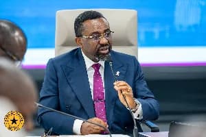 Dr. Mohammed Amin Adam, Ghana's Minister for Finance, is seen speaking into a microphone at a formal engagement. He is wearing a navy blue suit with a purple patterned tie and glasses, seated in a grey high-back chair.