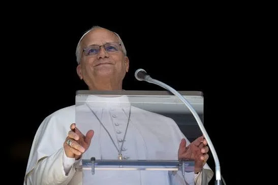 A photograph of Pope Leo standing at a transparent lectern with a microphone, looking upward with a slight smile. He is wearing traditional white papal attire, including a zucchetto, spectacles, and a pectoral cross against a solid black background.
