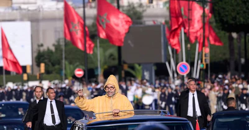 King Mohammed VI of Morocco waves to a crowd from an open-top vehicle during a public event. He is wearing a traditional yellow hooded djellaba and is flanked by security personnel in black suits, with several red Moroccan flags featuring the green star visible in the background.