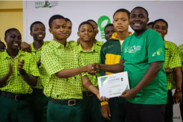 A group of Ghanaian students from GHANASS are pictured receiving a certificate of participation at an awards ceremony. A man in a green t-shirt shakes hands with a lead student while others in their school uniforms look on and smile in front of a YAFAT backdrop.
