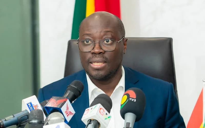 Dr. Cassiel Ato Forson, a prominent Ghanaian politician, is seen speaking at a press conference. He is dressed in a blue suit and glasses, positioned behind a cluster of microphones from major media outlets such as TV3 and Joy Business.