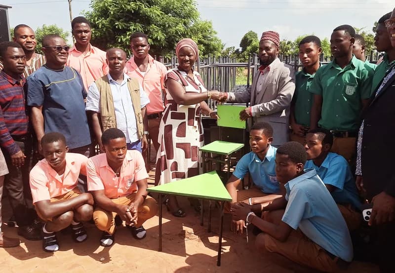 Techiman North MP Elizabeth Ofosu-Adjare is seen shaking hands with an official while presenting sets of green school desks and chairs. A large group of students in various uniforms and local representatives are gathered outdoors to witness the donation ceremony.