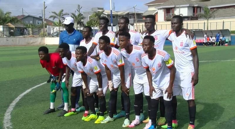 A Ghanaian football team poses for a group photograph on an artificial turf pitch. The players are dressed in white jerseys with orange and red details, while a coach or official in a blue shirt and white cap stands behind them. The background features residential buildings and a team dugout, typical of an urban Ghanaian sports setting.