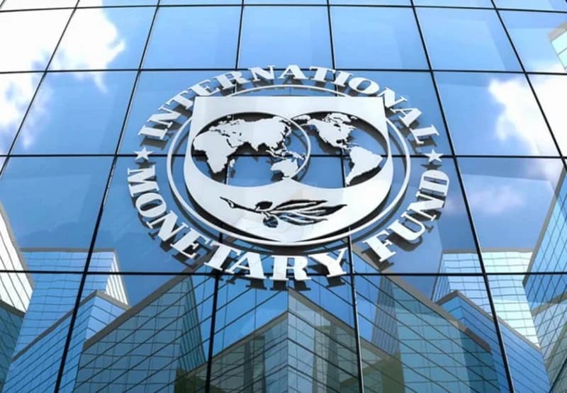 The image features the silver emblem of the International Monetary Fund (IMF) prominently displayed on the glass exterior of a modern skyscraper. The building's windows reflect surrounding architecture and a clear blue sky, emphasizing a corporate and global financial atmosphere.