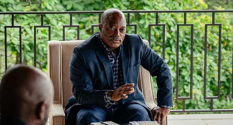 Former Democratic Republic of Congo President Joseph Kabila is depicted sitting in a chair on a balcony, engaged in conversation. He is dressed in a dark blazer and a plaid shirt, gesturing with his hand while looking toward another person whose head is partially visible in the foreground.