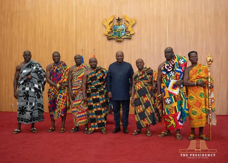 President John Mahama stands at the center of a group of traditional Ghanaian chiefs in a formal reception room, likely at Jubilee House. The chiefs are dressed in vibrant Kente cloths and traditional sandals, while the President wears a dark blue suit, positioned beneath the Coat of Arms of Ghana.
