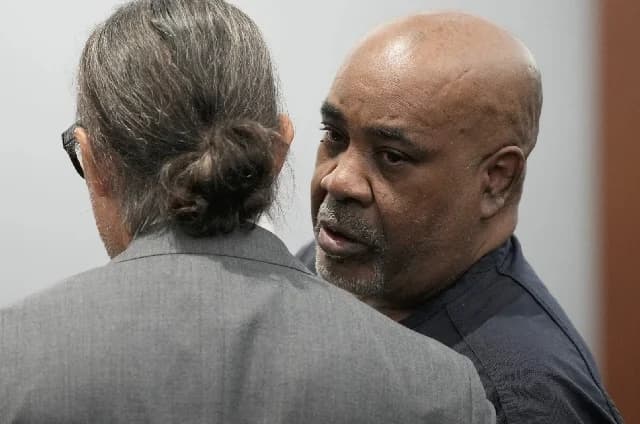 Duane 'Keffe D' Davis is captured in a courtroom setting, looking toward another individual who has their back to the camera. Davis, with a shaved head and grey goatee, appears to be listening or waiting during legal proceedings related to the Tupac Shakur murder case.