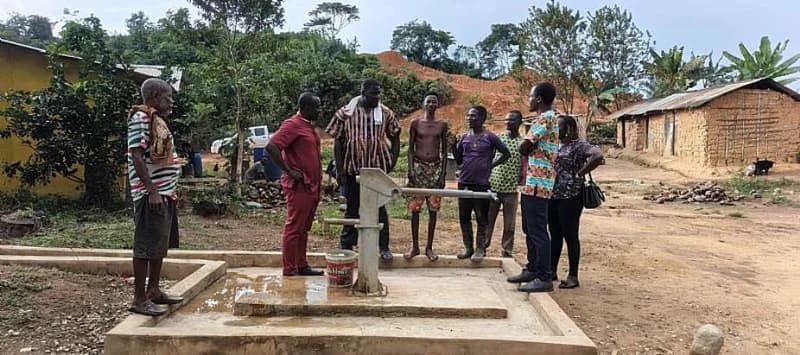 A group of people, including community members and officials, are gathered around a manual water pump on a concrete platform in a rural setting. The scene depicts a site visit or sensitization meeting involving infrastructure development within a Ghanaian district.