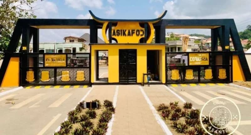 The image shows the ornate yellow and black entrance gate of Obuasi Senior High Technical School (SHTS) in Ghana. The architecture features a central security booth topped with a Ghanaian traditional stool motif, with paved driveways and landscaped plant beds in the foreground.