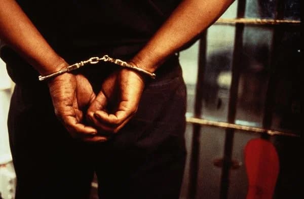 A close-up photograph shows a person with their hands handcuffed behind their back. The subject stands in front of metal cell bars, clearly indicating a jail or prison environment.