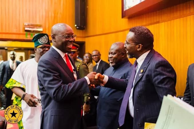 Governor of the Bank of Ghana, Dr. Ernest Addison, is seen shaking hands and smiling with another official during a formal gathering. The setting features wood-paneled walls and includes various dignitaries, one wearing traditional Ghanaian headgear and another in military attire.