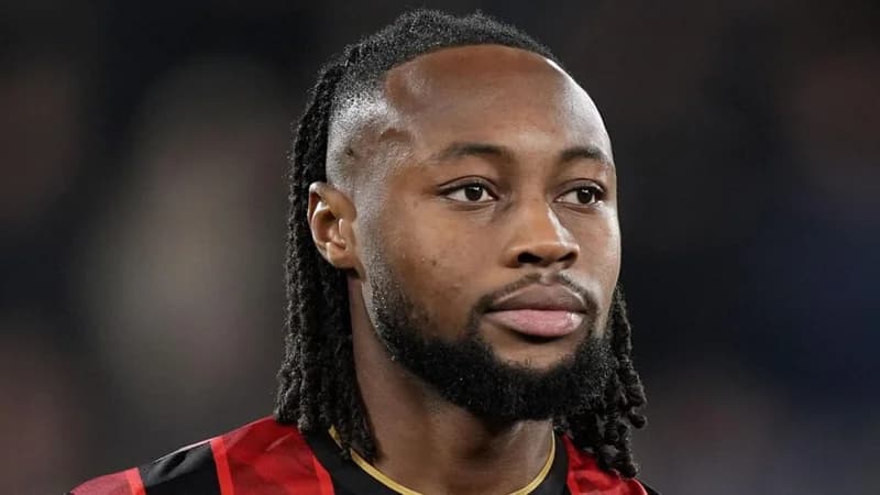 A close-up portrait of Ghanaian international footballer Antoine Semenyo. He is seen wearing a red and black striped football jersey with a gold trim, looking towards the side with a neutral expression.