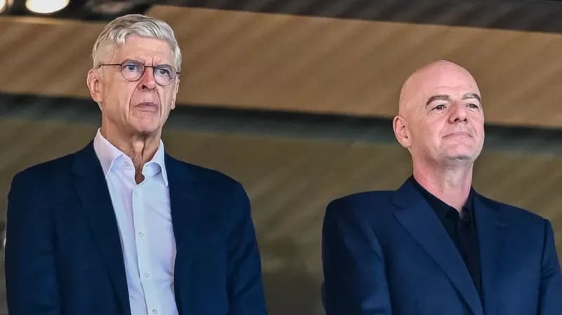 Arsène Wenger and Gianni Infantino are captured in a medium shot, standing together at what appears to be a football stadium or official event. Wenger, on the left, wears spectacles and a white shirt under a blue suit, while Infantino, on the right, wears a dark shirt with a matching blue suit.