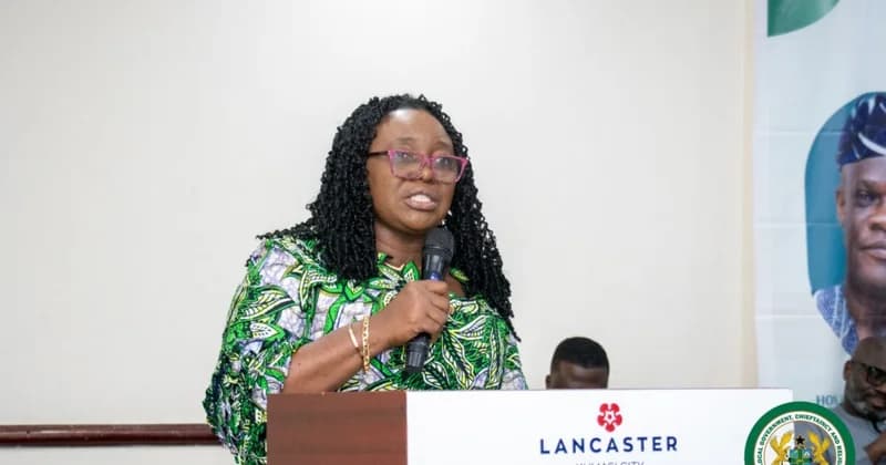 A woman with dark curly hair and pink-framed glasses is speaking into a microphone at a podium. She is wearing a green-patterned dress and appears to be delivering a speech at an event hosted at Lancaster Kumasi City, as indicated by the logo on the podium.