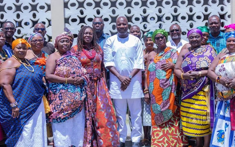 A group of Ghanaian Queen Mothers dressed in vibrant traditional kente fabrics and headwraps pose for a photograph with a male official in a white smock. The group is gathered in front of a white wall featuring a distinctive circular breeze block pattern.