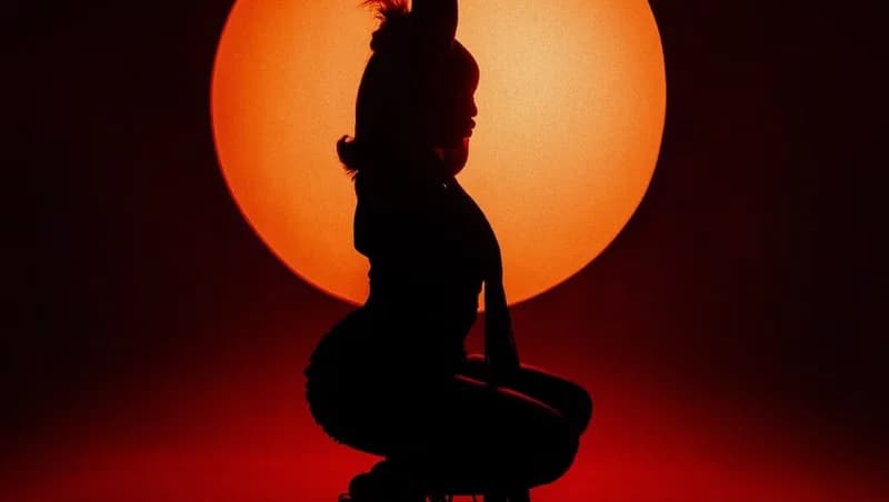 A silhouette of a person, likely the artist Gyakie, is captured in a crouching pose against a large, glowing orange circular light source. The background is a deep, moody red, creating a dramatic and artistic aesthetic typical of a music video still or promotional cover art.