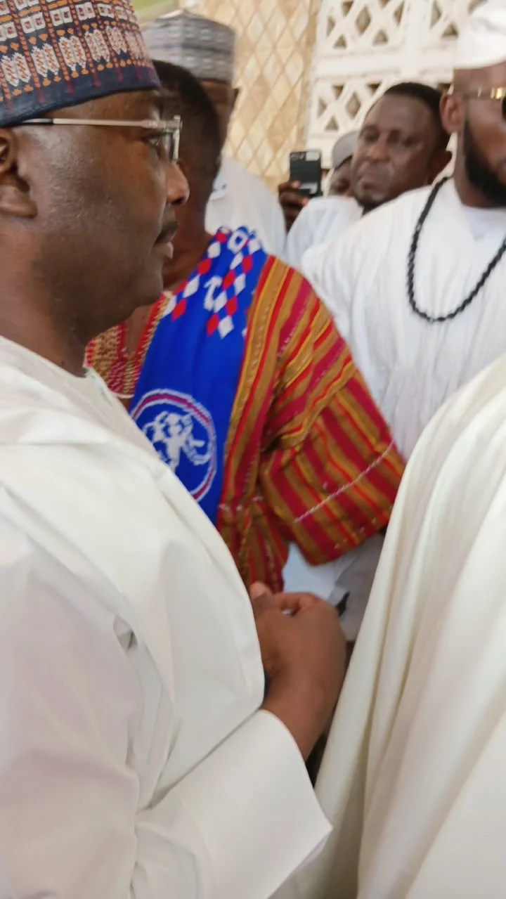 Dr. Mahamudu Bawumia is captured in a profile view while attending a public event, surrounded by several individuals in traditional attire. A person standing behind him is wearing a smock prominently featuring the New Patriotic Party (NPP) elephant logo, suggesting a political gathering or party function.