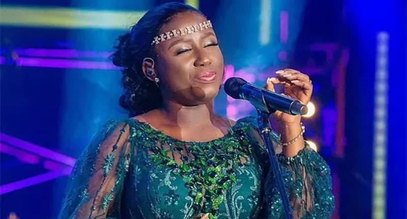 Ghanaian gospel singer Diana Hamilton is captured performing on stage, wearing an elegant green sequined dress and a decorative headband. She has her eyes closed in a soulful expression while singing into a microphone against a backdrop of purple and blue stage lights.