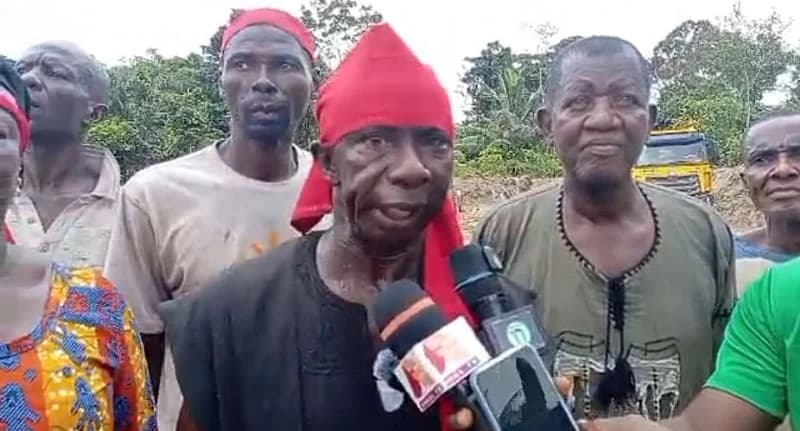 A group of residents, including farmers, are gathered in Assin Kumasi for a media address regarding local tensions. The central figure, wearing a red headscarf symbolizing distress or protest in Ghanaian culture, speaks into several microphones. In the background, a yellow truck and other community members wearing red headbands are visible against a backdrop of trees.