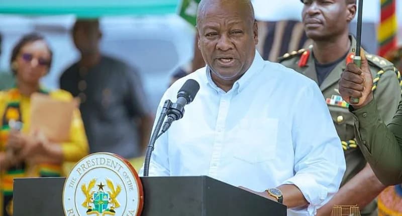 President John Dramani Mahama is pictured delivering an official address from behind a presidential podium. He is wearing a white shirt, while a military aide-de-camp stands behind him holding an umbrella during what appears to be an outdoor state function.